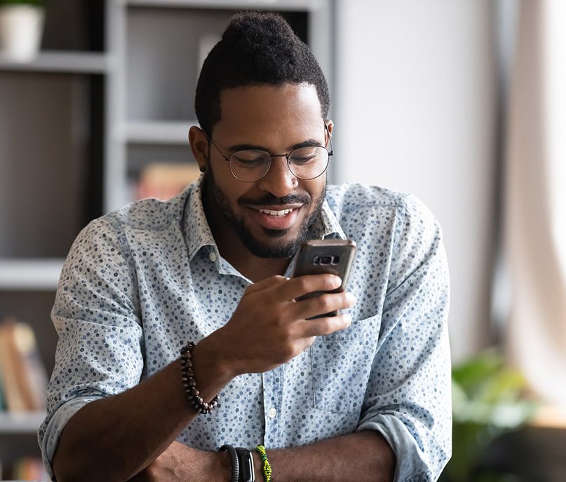 Feliz joven africano hombre de negocios estudiante profesional usuario mantenga smartphone moderno gadget mensaje de texto en la aplicación de redes sociales en línea sentarse en el escritorio de la oficina en casa utilizando tecnología de teléfono celular para navegar por la web